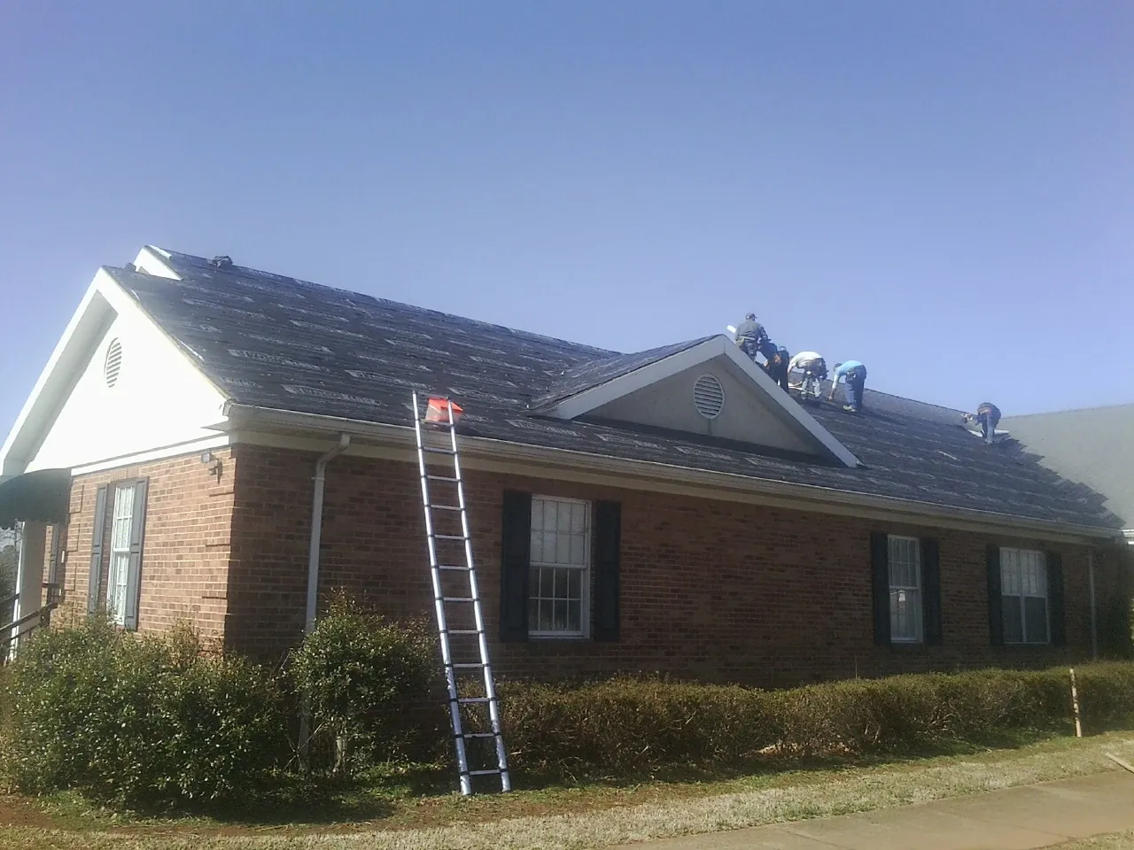 Crew tearing shingles off a brick ranch roof with a ladder set — Emergency Roof Repair work by our crew in Nemacolin
