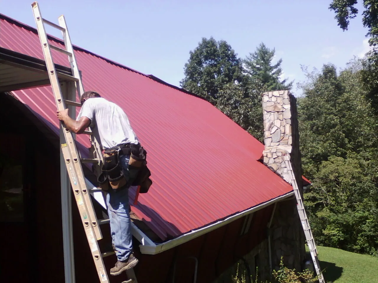 Roofer on a ladder beside a red metal roof and stone chimney — Soffit & Fascia Repair by Roofing Brothers in Nemacolin